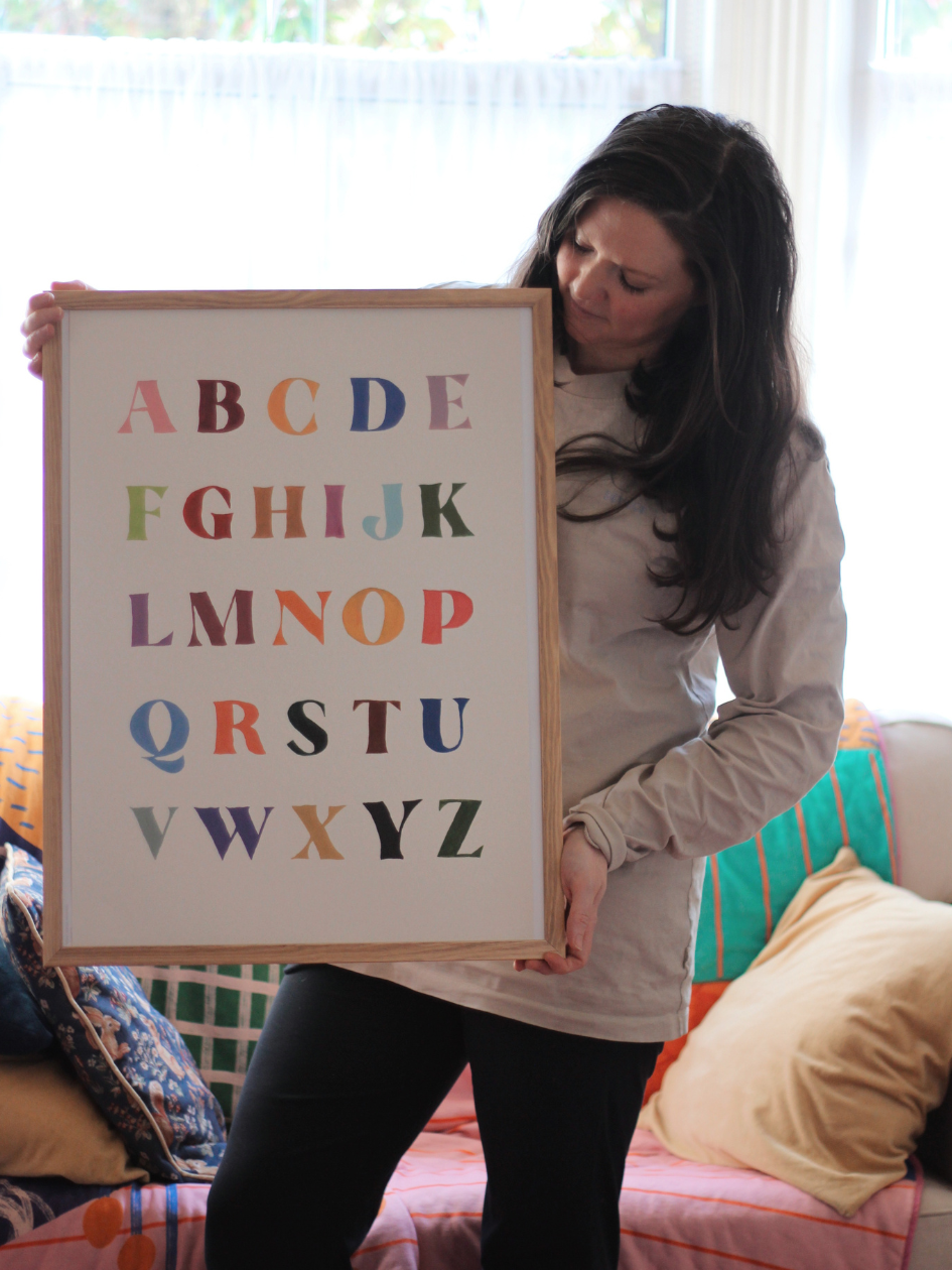 Woman holding a framed alphabet poster in a home setting